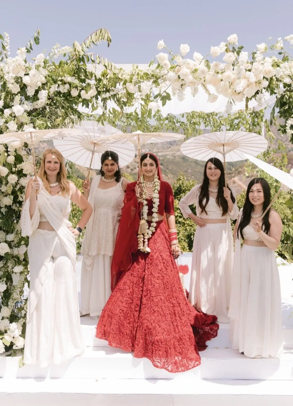 Bride in red with her bridesmaids holding parasols under a white floral mandap
