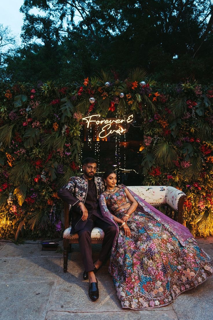 Couple at their sangeet under a floral wall and neon sign